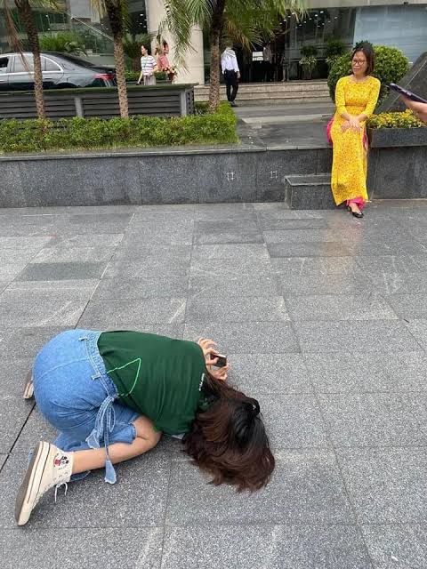 A photographer crouching low on a paved plaza to take a portrait of a woman in a yellow ao dai