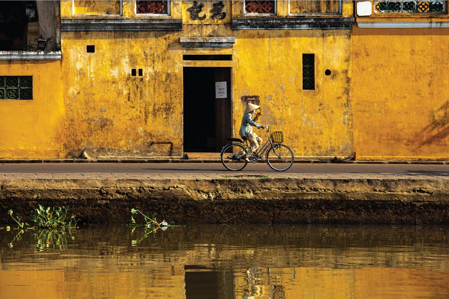 A woman in a conical hat cycling past yellow shophouse walls by the river in Hoi An old town