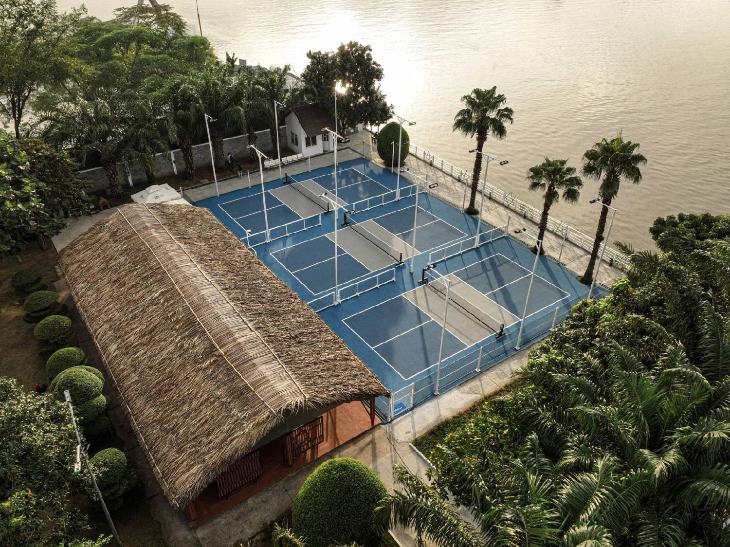 Aerial view of three blue pickleball courts beside a river with a thatched clubhouse in Saigon