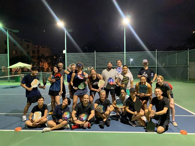 A mixed group of visitors and local players posing with paddles on a floodlit pickleball court at night in Vietnam